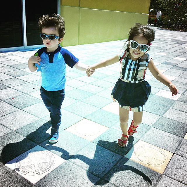 Two stylish children holding hands walking on a sunny paved area.