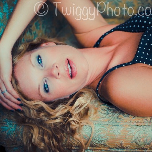 A beautiful blonde woman with blue eyes poses intimately on a vintage couch in a soft, dreamy style.