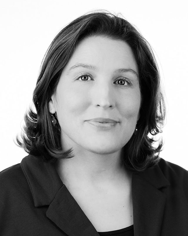 Professional headshot of a confident woman in a dark suit against a white background.