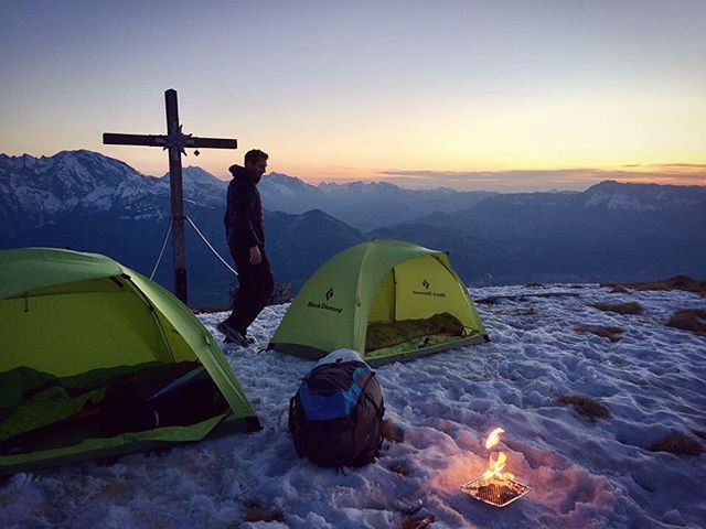 A man camps on a snowy mountaintop, with tents and a fire under a vibrant sunrise. 