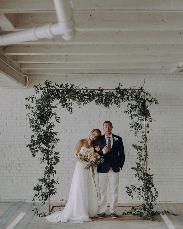 A happy couple poses in front of a greenery-covered wedding arch on their special day.