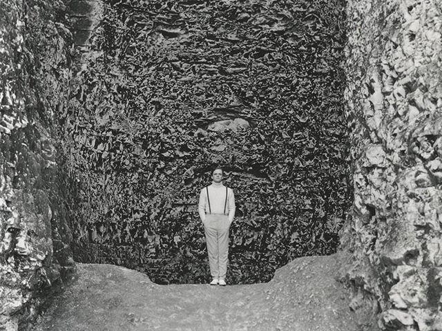 A man stands within a large, textured cave, captured in a monochrome shot. 