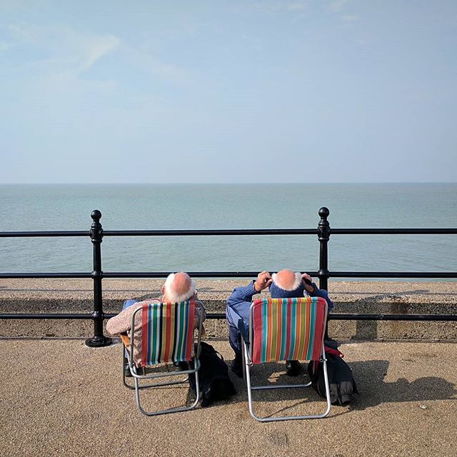 An elderly couple relaxes in deck chairs by the sea on a sunny day, enjoying the calm ocean view.