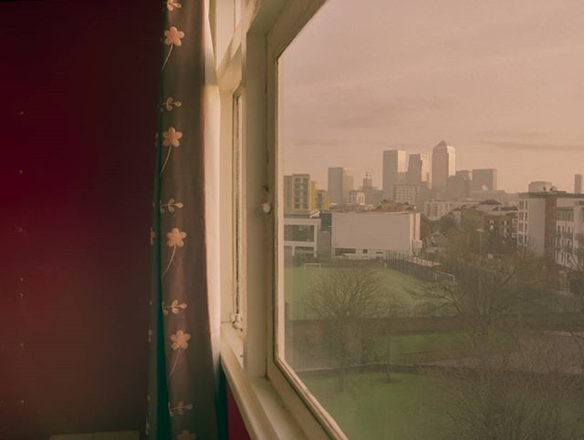 A view of a cityscape through a window with a floral curtain. The buildings in the skyline are shrouded in a soft haze.