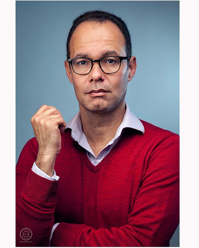 Professional portrait of a man wearing glasses and a red sweater against a blue backdrop.