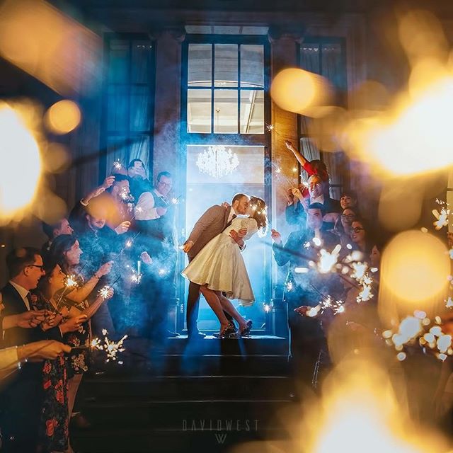 A bride and groom kiss at their wedding exit, surrounded by guests holding sparklers.
