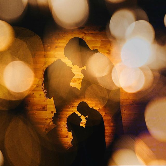 Silhouetted couple embrace against a wooden backdrop, softly blurred by bokeh lights, suggesting intimacy and connection.