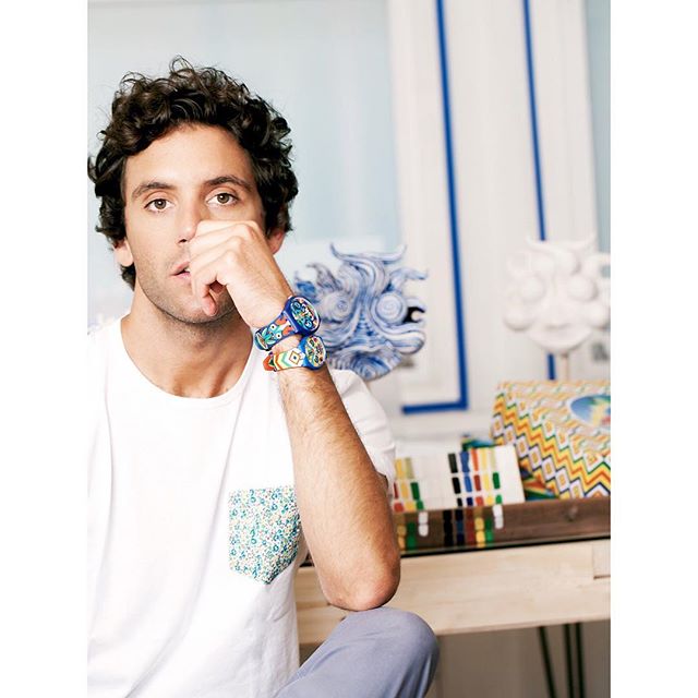A man sits indoors wearing two colorful watches, hand to his face in a contemplative pose.