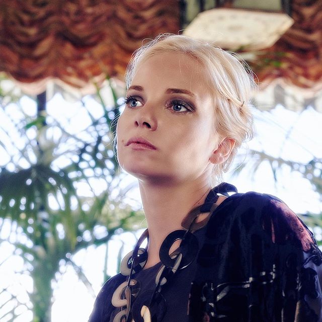 A blonde woman with blue eyes poses indoors with natural light, showcasing a contemplative and serious mood.