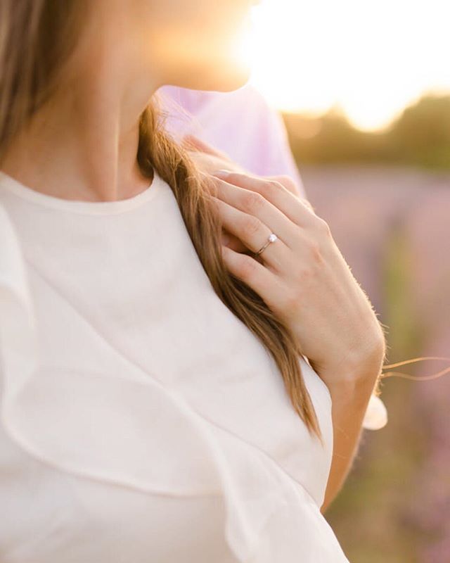 Close-up of a woman's hand wearing an engagement ring, with a couple in the background at sunset.