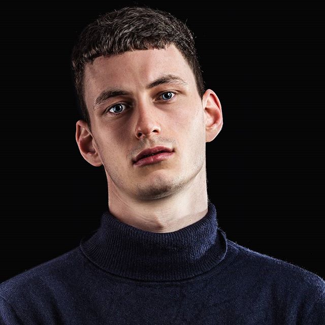 A man with blue eyes and short hair poses against a dark background in a portrait.