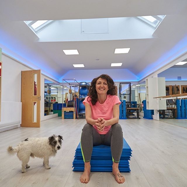 A smiling woman sits on a stack of exercise mats next to her dog in a Pilates studio.