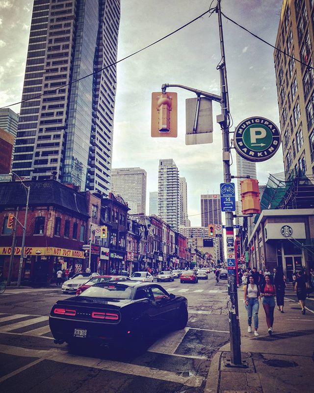 A bustling city street scene featuring pedestrians, vehicles, and towering buildings under a cloudy sky.
