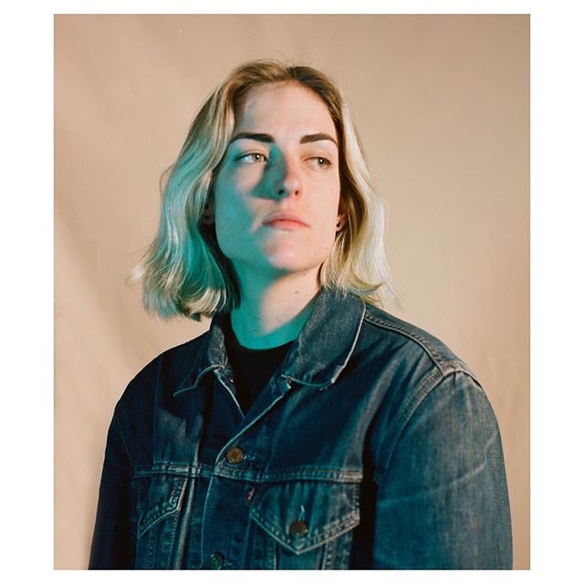 A woman in a denim jacket stares pensively in a studio, bathed in soft, cool-toned lighting.