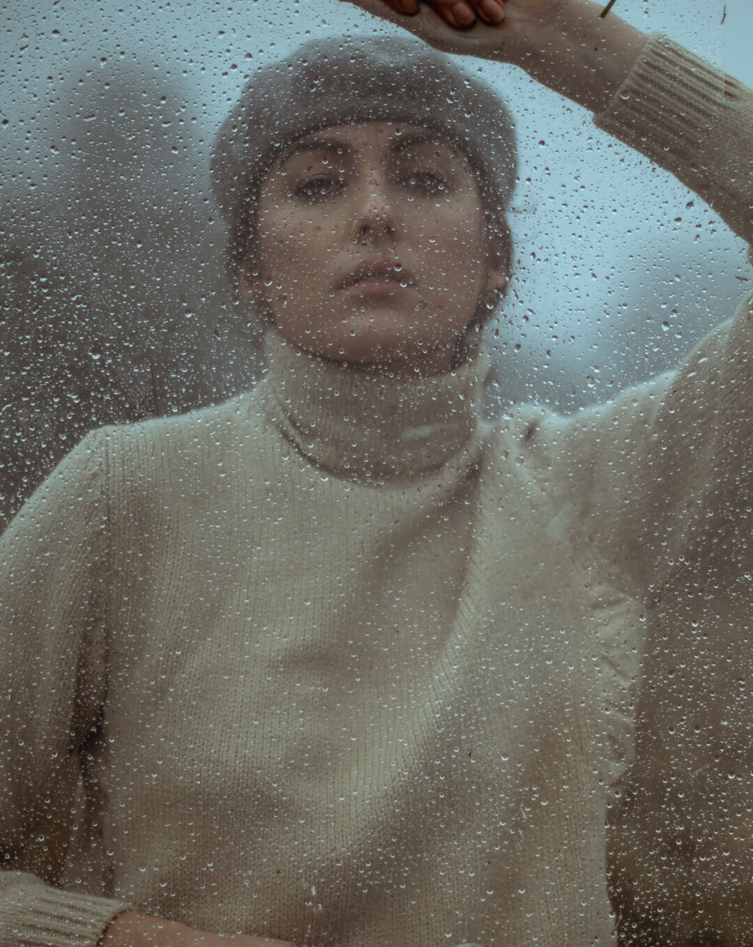 A woman in a beret and turtleneck sweater poses behind a rain-streaked window.