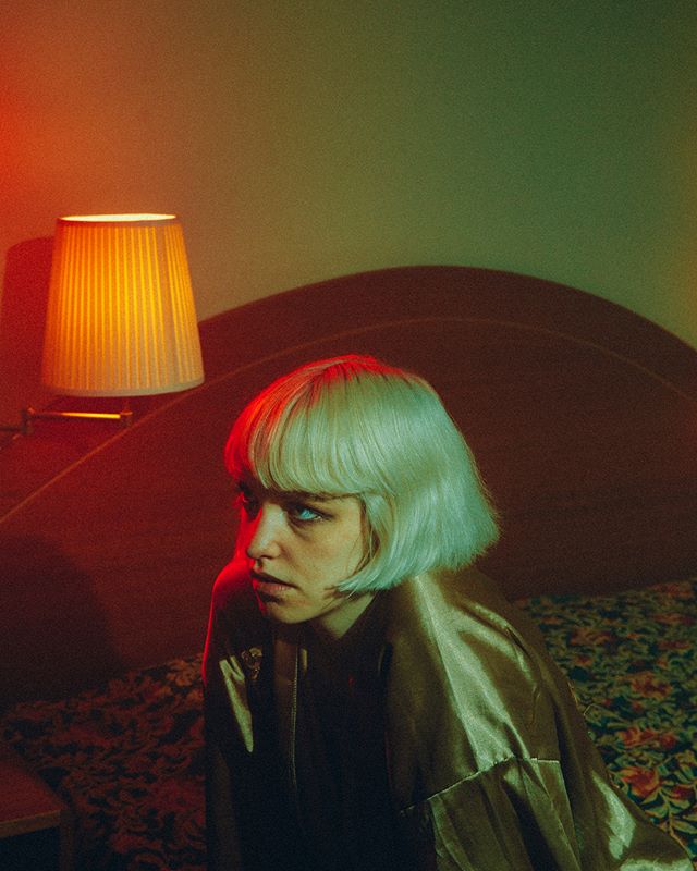 A woman with a blonde bob sits on a bed with red lighting in a moody setting.