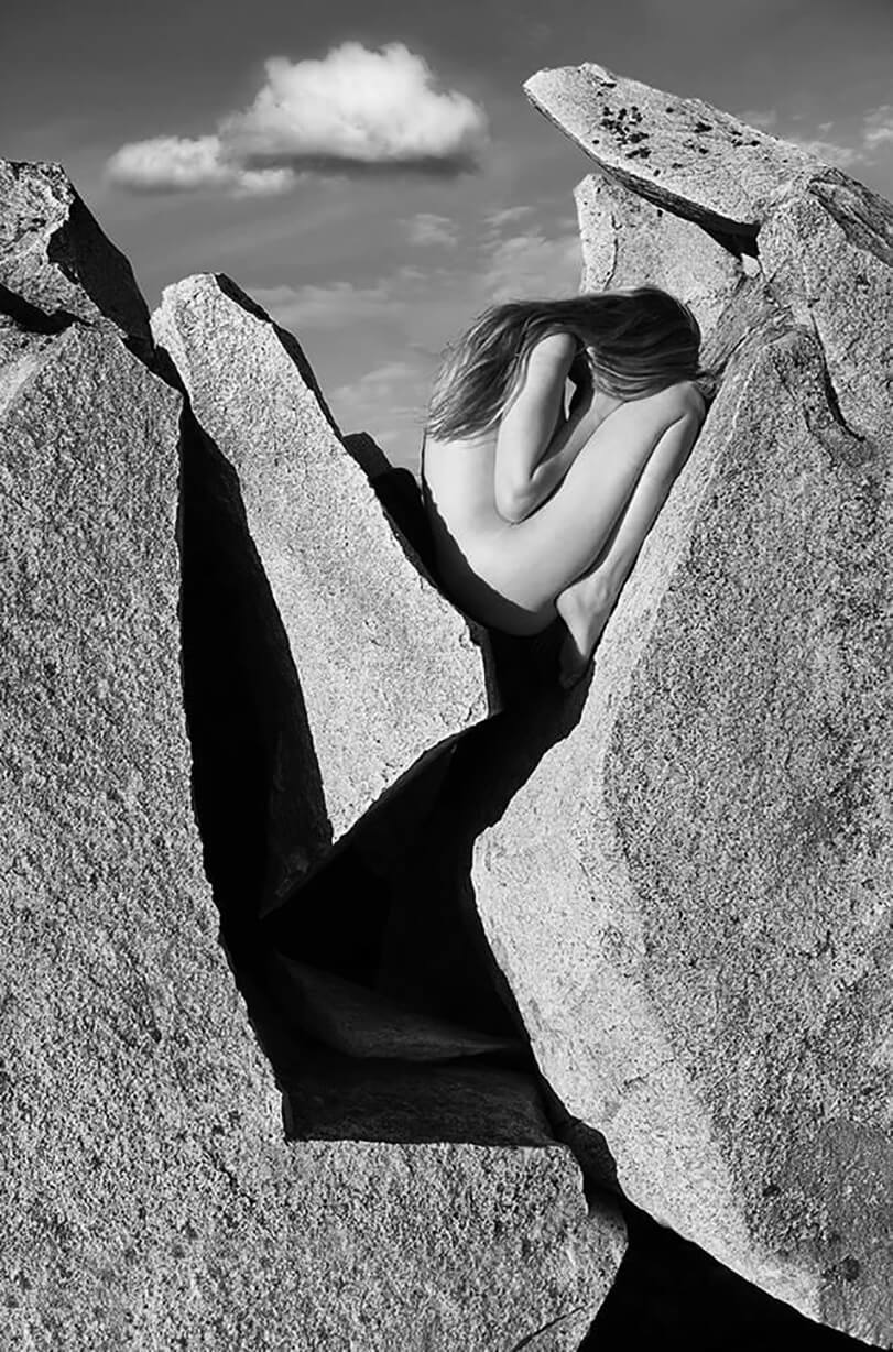 A nude woman sits nestled among rocks in a black and white artful photograph, evoking introspection and connection with nature.