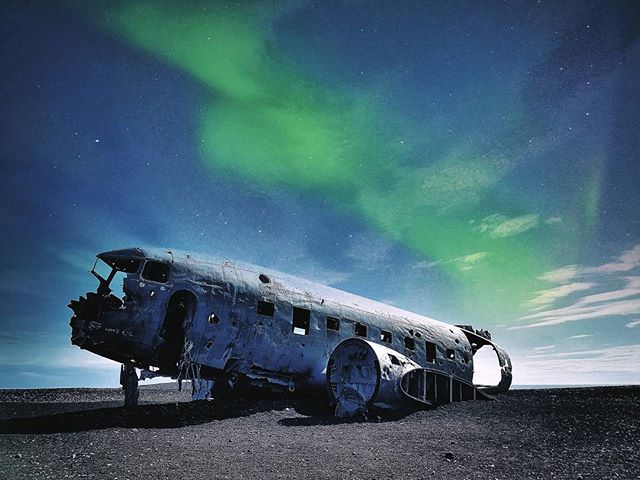 An airplane wreck rests on a black sand beach under a sky illuminated by the green aurora borealis at night in Iceland.