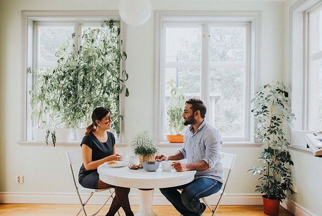 A couple enjoys a breakfast at a round table near a window filled with plants in their home.