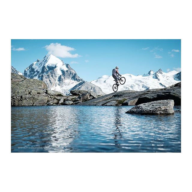 A cyclist performs a stunt on a mountain bike with a scenic mountain and lake backdrop.