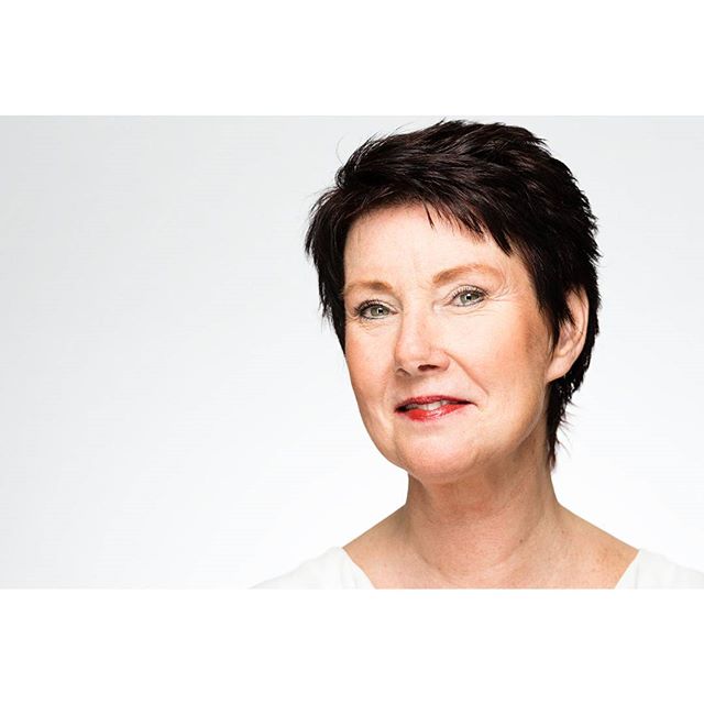 A mature woman with short dark hair poses for a professional headshot in a studio setting.