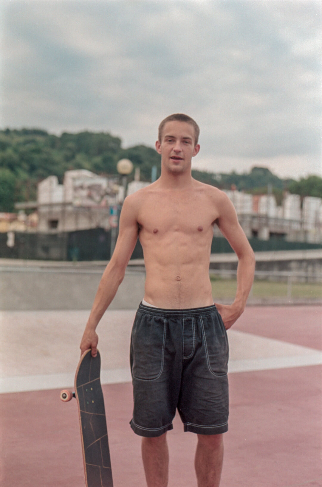 A shirtless man in shorts holds a skateboard at the skatepark.