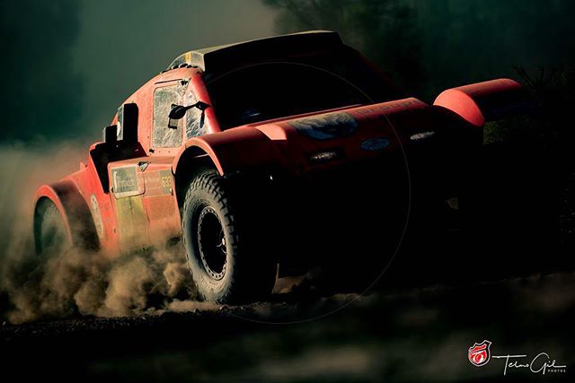 A red rally car speeds across an off-road track, kicking up dust in a dynamic action shot.