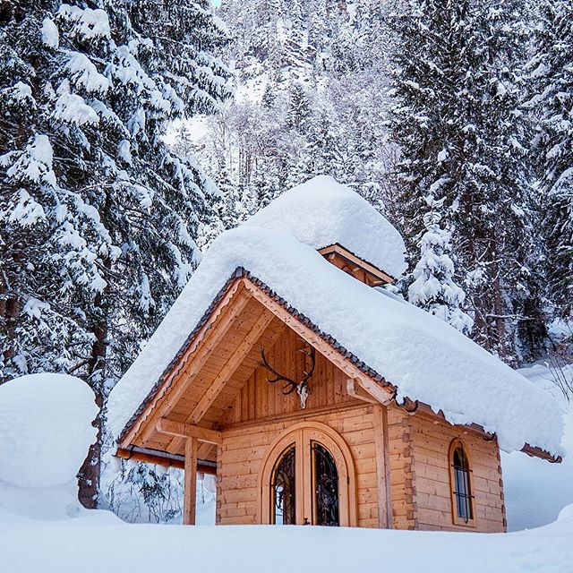 A cozy cabin is nestled in a snowy forest, its roof covered in fresh snow. Deer antlers decorate the entrance.