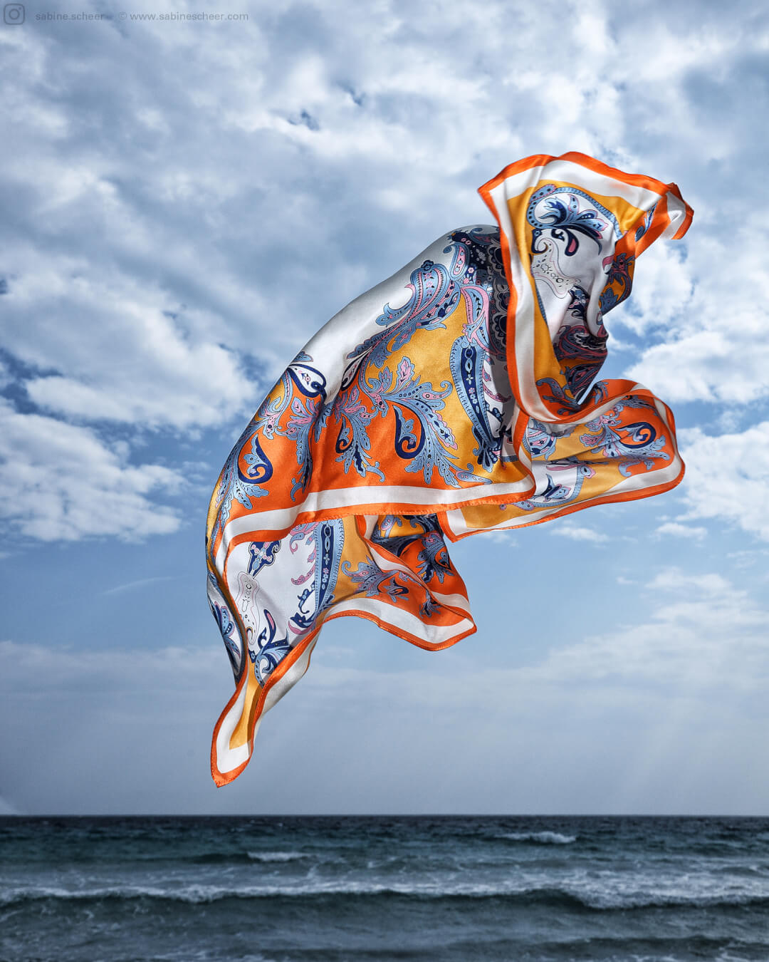 A brightly colored silk scarf floats in the air above a calm ocean under a partly cloudy sky.