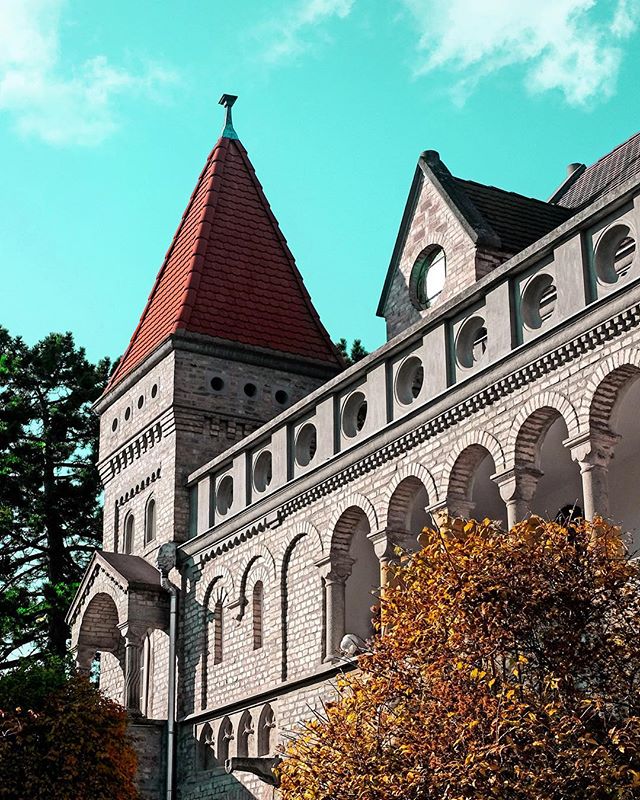 Architectural shot of a castle tower and facade with a red roof against a partly cloudy turquoise sky.