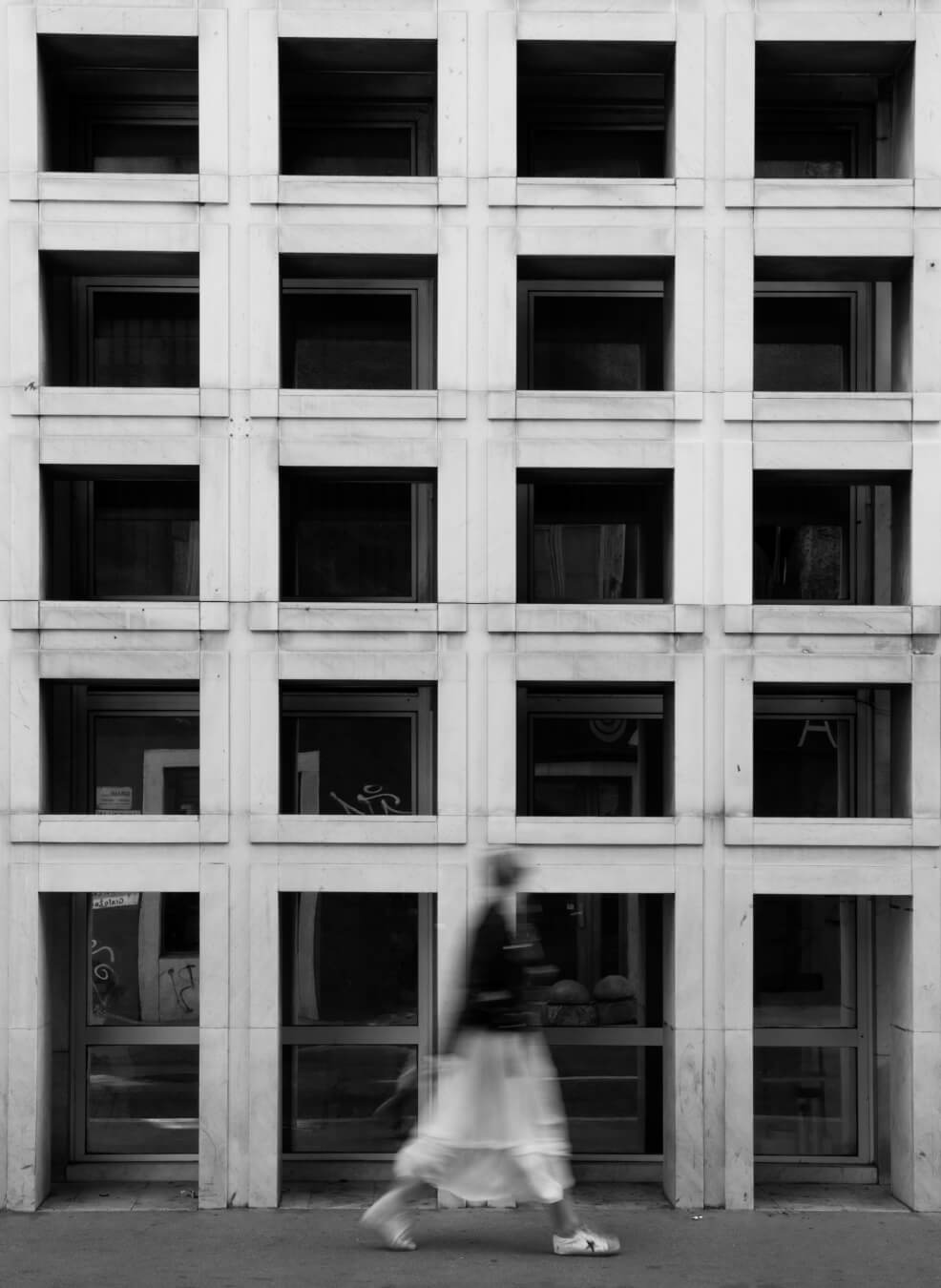 A blurred figure walks past a building's facade with repeating window patterns in black and white.