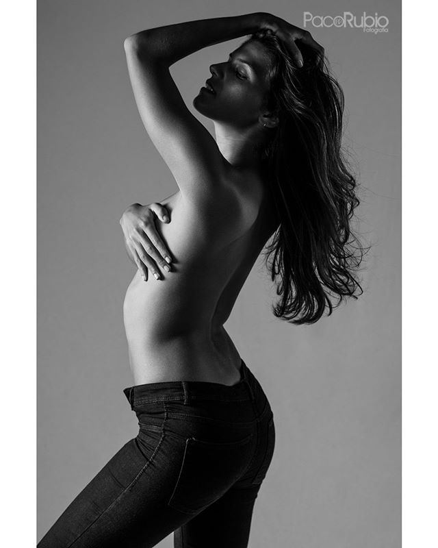 Black and white portrait of a topless woman in jeans, hand covering her breast, in a studio.