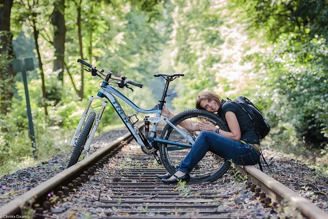 A woman relaxes with her Trek mountain bike on the railroad tracks amidst a lush green forest.