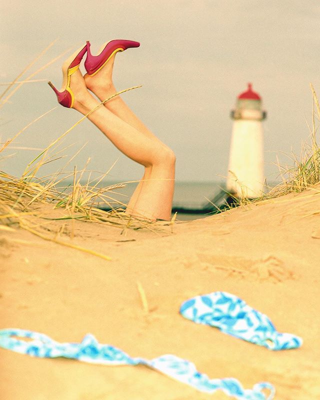 A woman's legs wearing heels are sticking out of the sand, with a swimsuit and lighthouse visible.