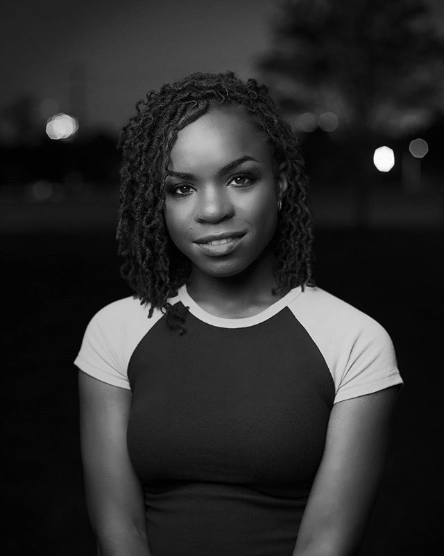 A confident black woman poses in a black and white portrait with a blurred background at night.