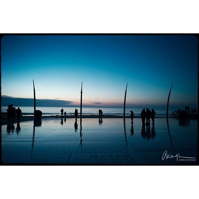 Silhouetted people stroll along a reflective waterfront at dusk.