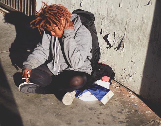 A person with dreadlocks sits against a wall in an urban setting, appearing contemplative and moody.