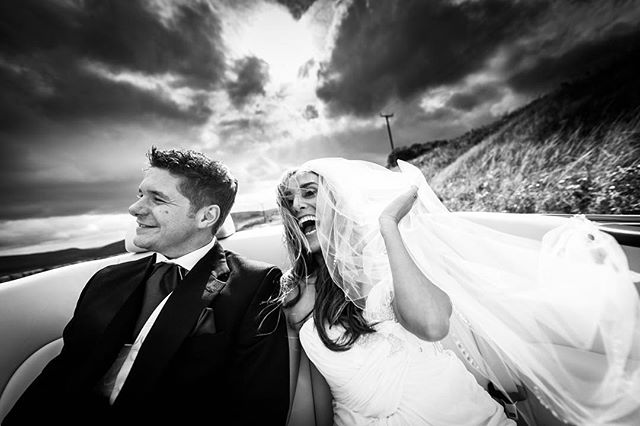 A happy bride and groom ride in a car, the bride's veil catching the wind, celebrating their wedding day.
