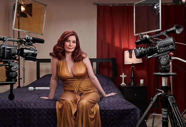 A mature woman poses on a bed in a film studio, surrounded by cameras and lighting equipment.