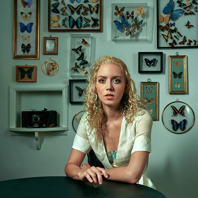 A portrait of a curly haired woman in front of a wall decorated with framed butterflies.