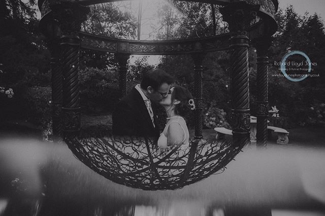 A bride and groom share a kiss within a gazebo, captured in a romantic, black-and-white wedding photograph.