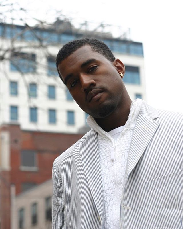 A medium shot of Kanye West wearing a striped suit jacket in an urban setting. 