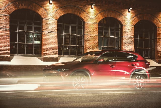 A red Mazda CX-5 speeds along a city street at night, captured with a long exposure to emphasize movement.