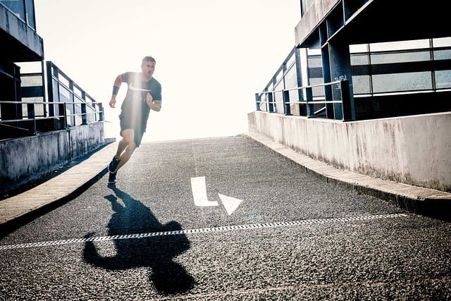 A determined athlete runs uphill on a city street, shadowed by urban buildings and bright sunlight.