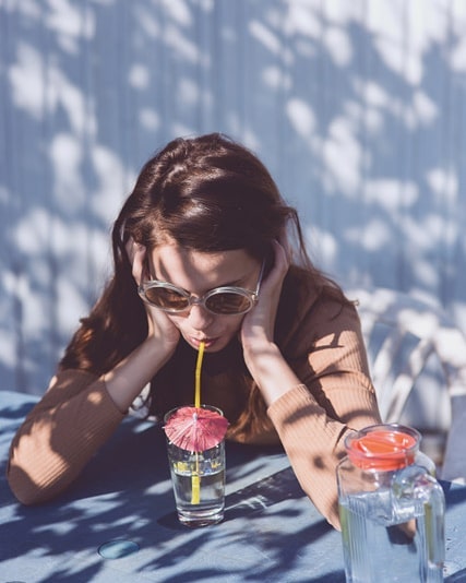 A woman sips a drink with an umbrella straw at an outdoor table in dappled sunlight.