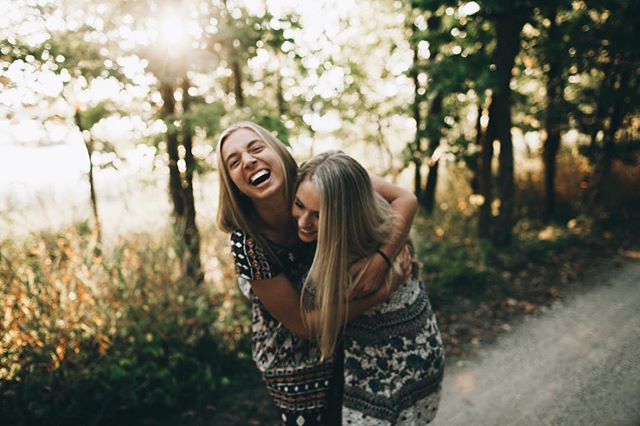 Two women friends are laughing and hugging outdoors in a sunny forest setting.