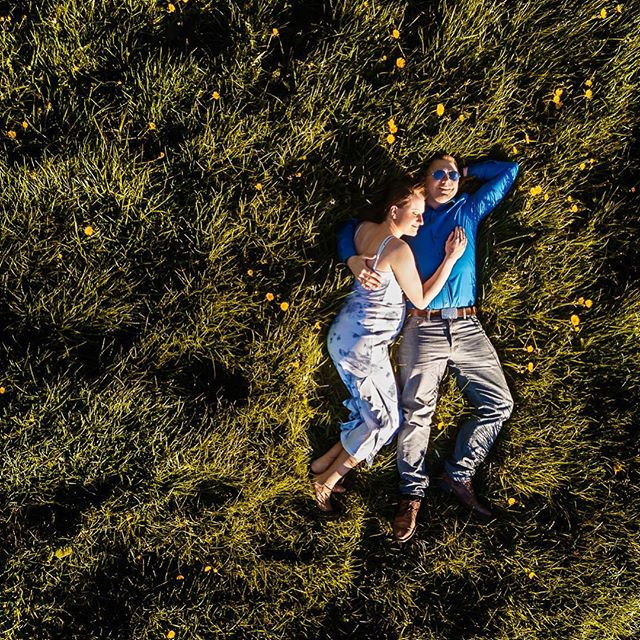A couple lies in a grassy field, embracing, captured from above. Serene and intimate moment in nature.