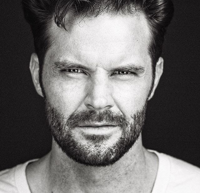 A black and white close-up portrait of a serious man with a beard against a dark background.