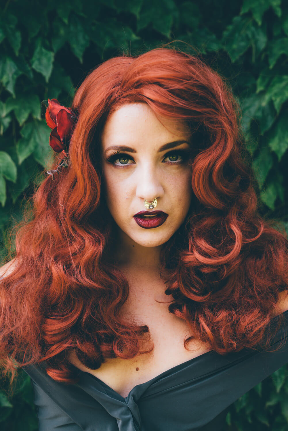A striking woman with red hair, a septum piercing, and red lipstick poses against a lush green backdrop.