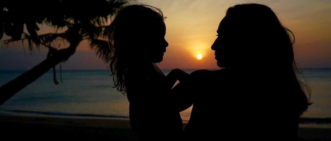 A silhouette shows a mother holding her child on a beach at sunset. Warm tones and a loving mood evoke a peaceful scene.